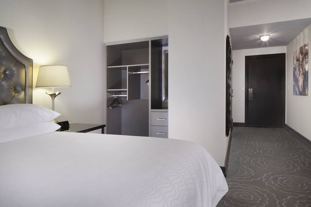 A hotel room with a bed, a nightstand, a lamp, a wardrobe, and a door leading to another room.