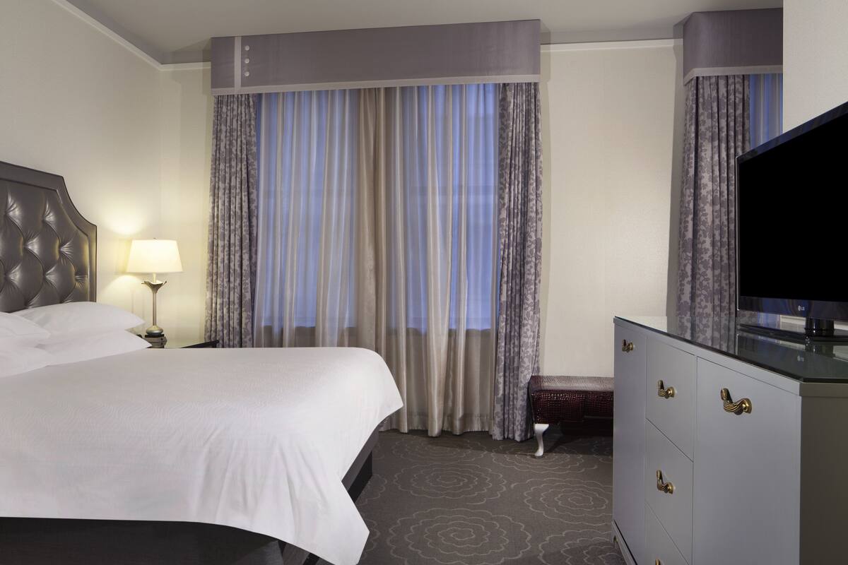 A hotel room with a large bed, a bedside table with a lamp, a dresser with drawers, and a television.