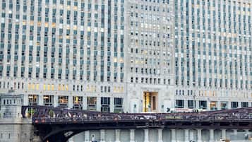 A large building with a river in front, a bridge, and people relaxing on chairs.
