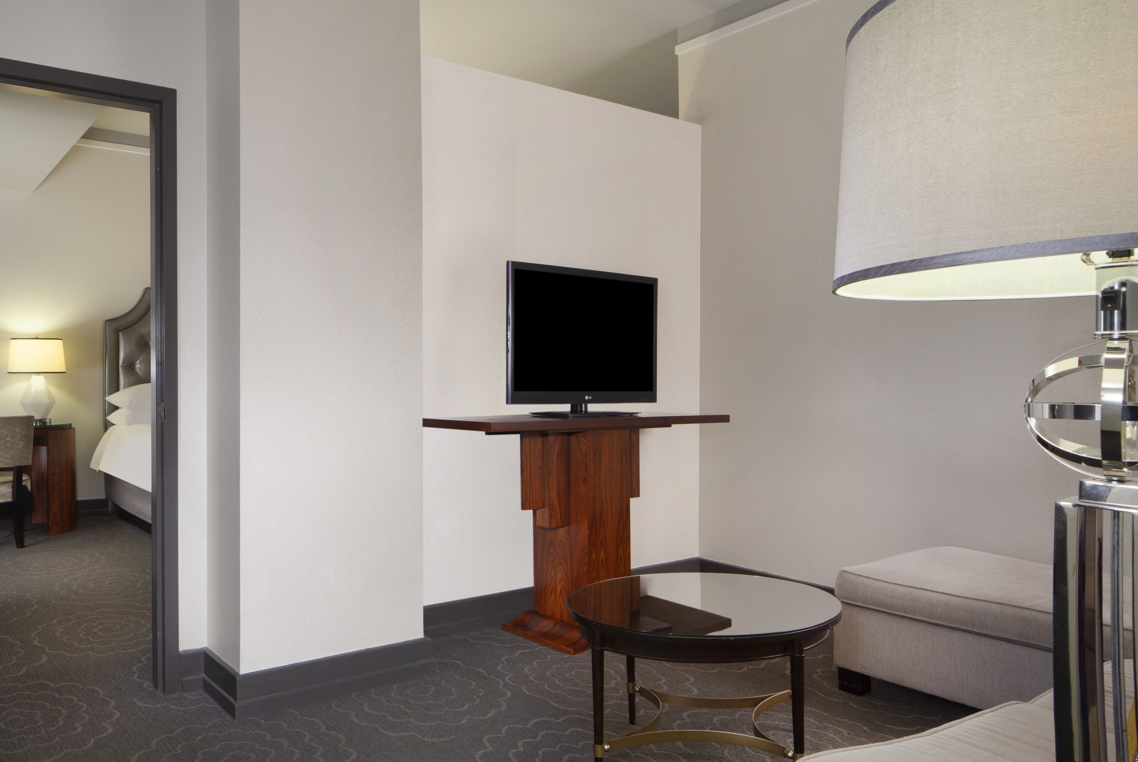 A hotel room with a flat-screen TV on a wooden stand, a glass table, a sofa, and a lamp.