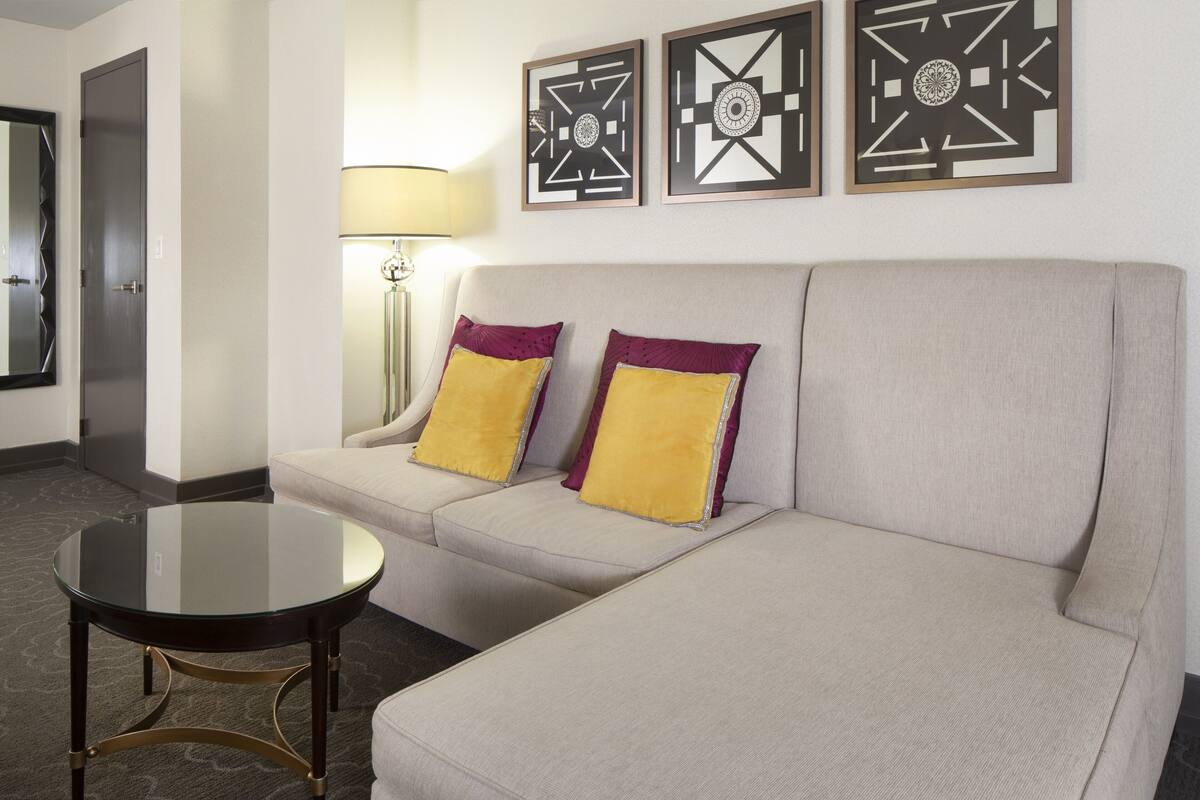 A modern hotel room with a beige sofa, two yellow and purple pillows, a glass coffee table, and three geometric wall art pieces.