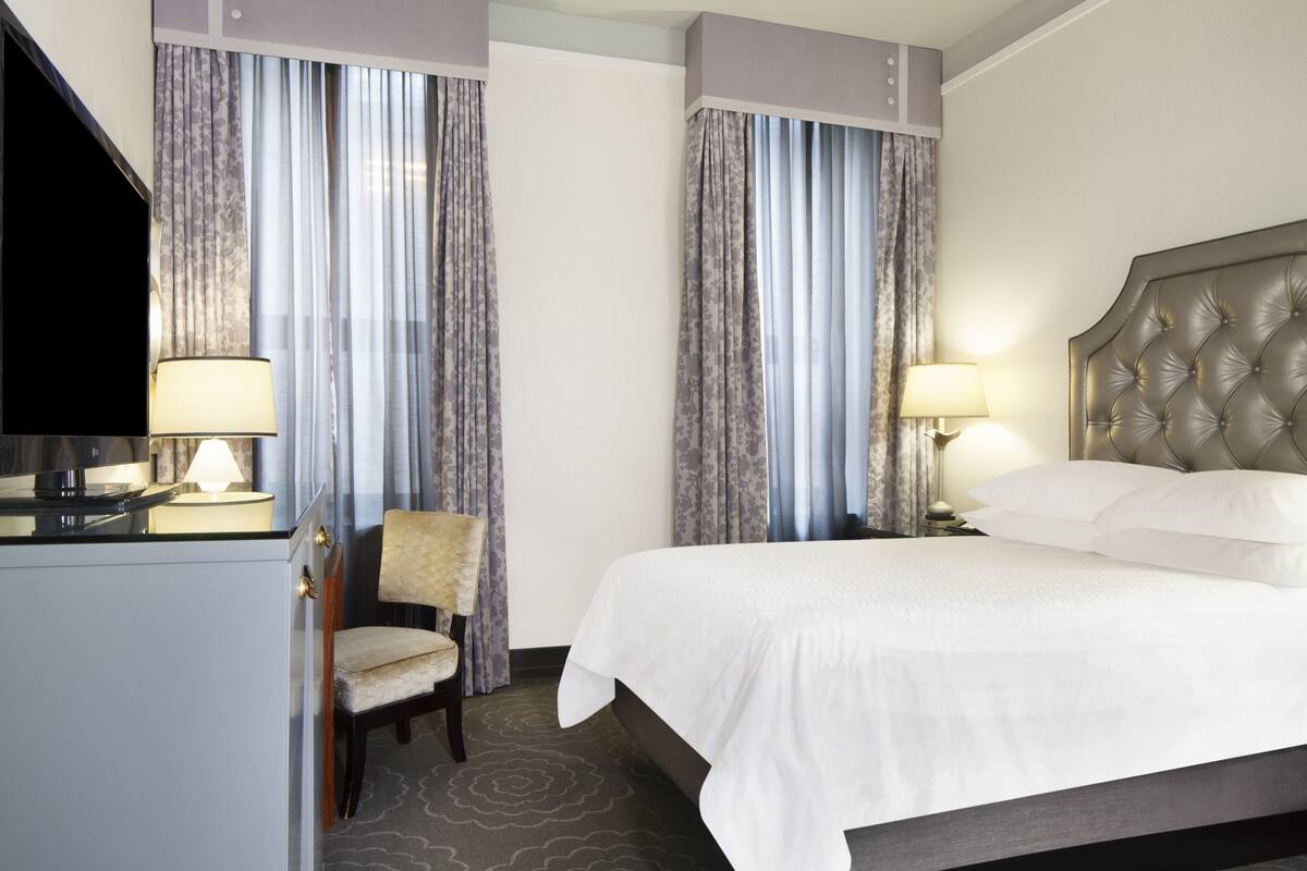 A hotel room with a bed, two bedside lamps, a chair, a TV, and curtains.