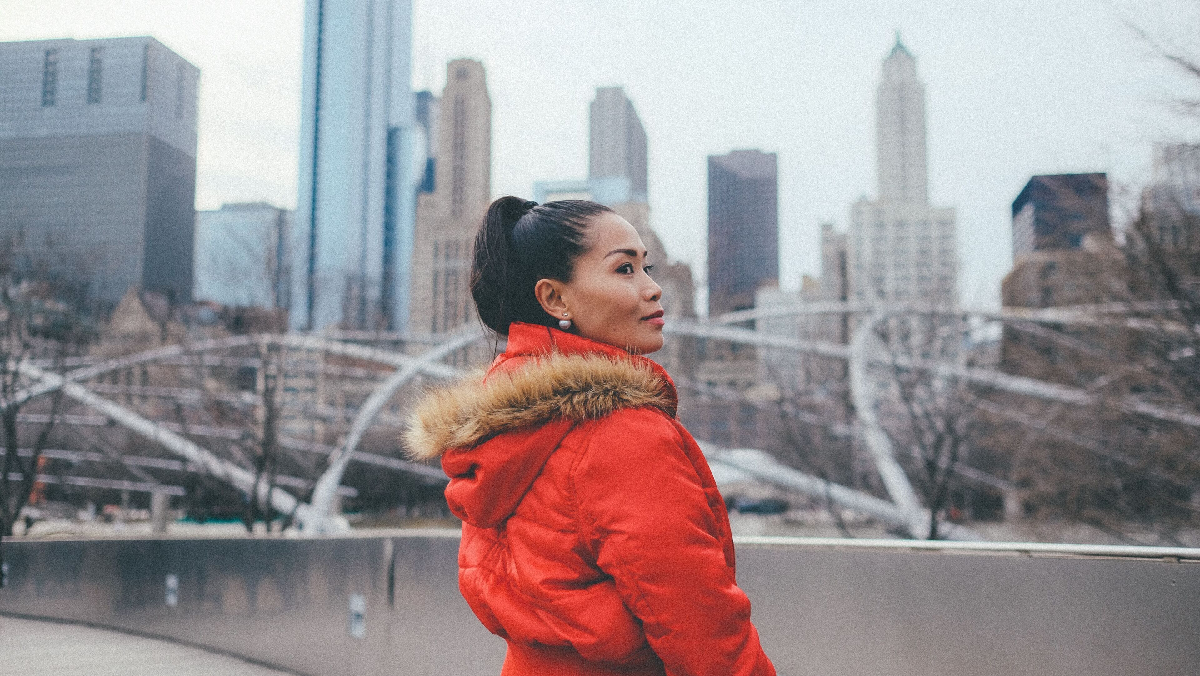 Una persona con una chaqueta roja y capucha de piel de pie frente a un horizonte urbano con edificios altos y un puente.