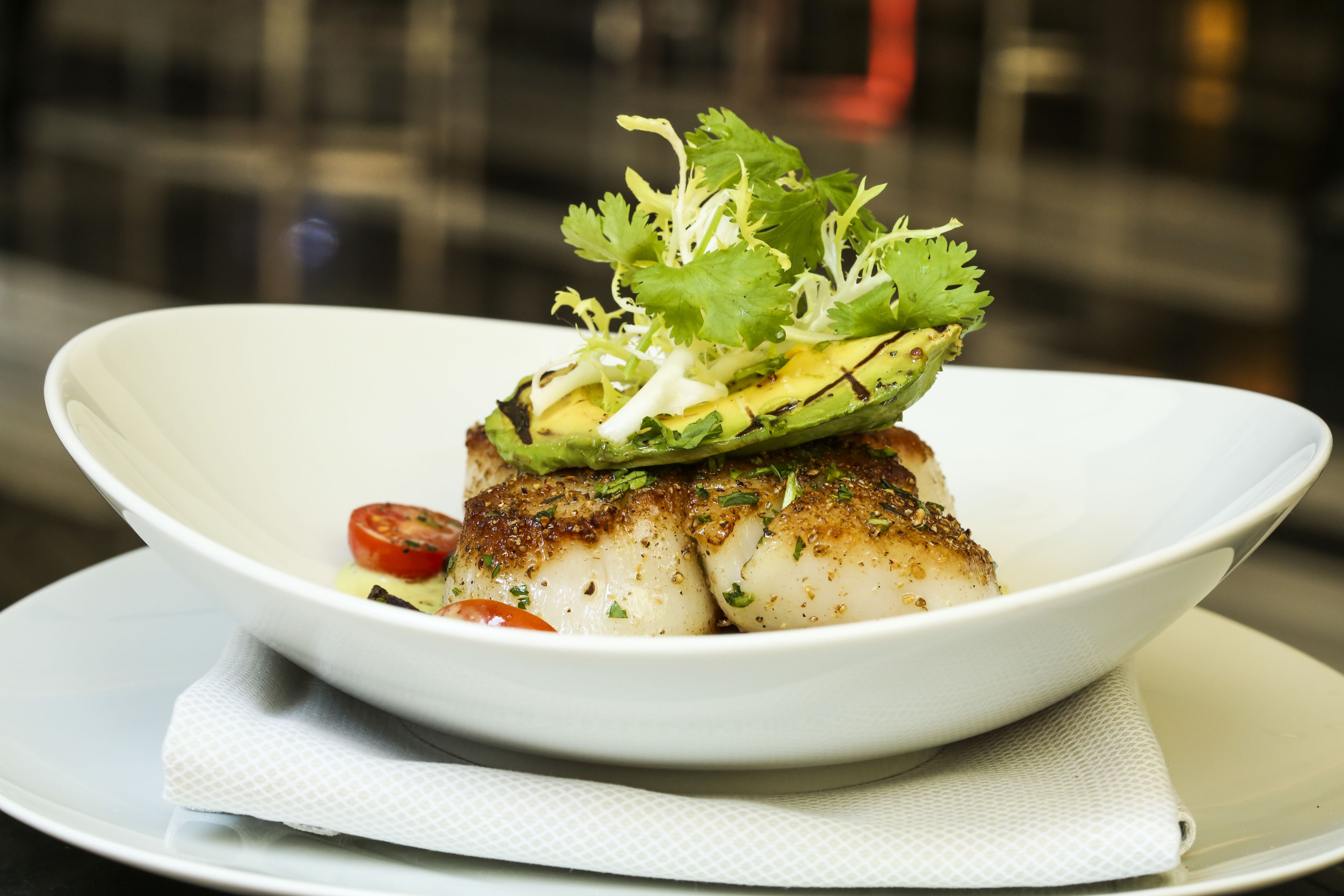 Un tazón blanco con un trozo de pescado a la parrilla cubierto de aguacate, cilantro y una rodaja de tomate.