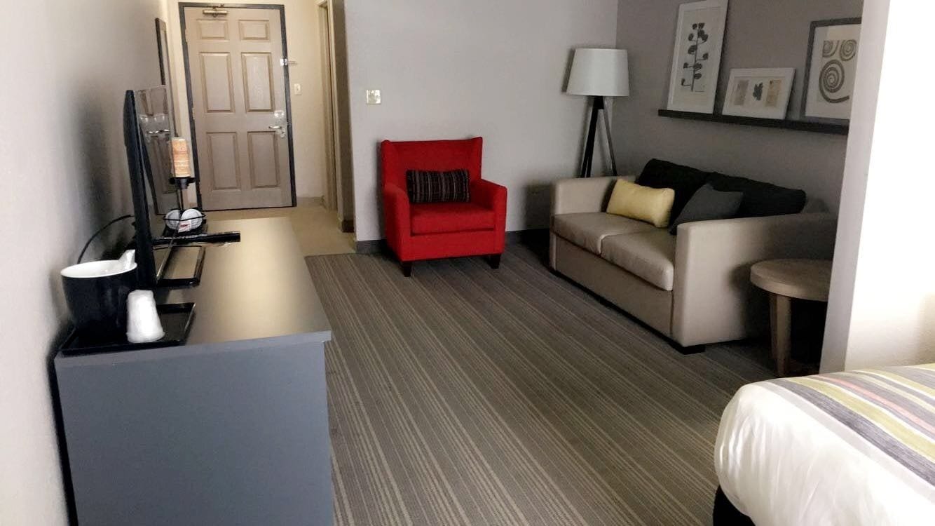 A hotel room with a bed, a red armchair, a sofa, a desk with a lamp, and framed pictures on the wall.