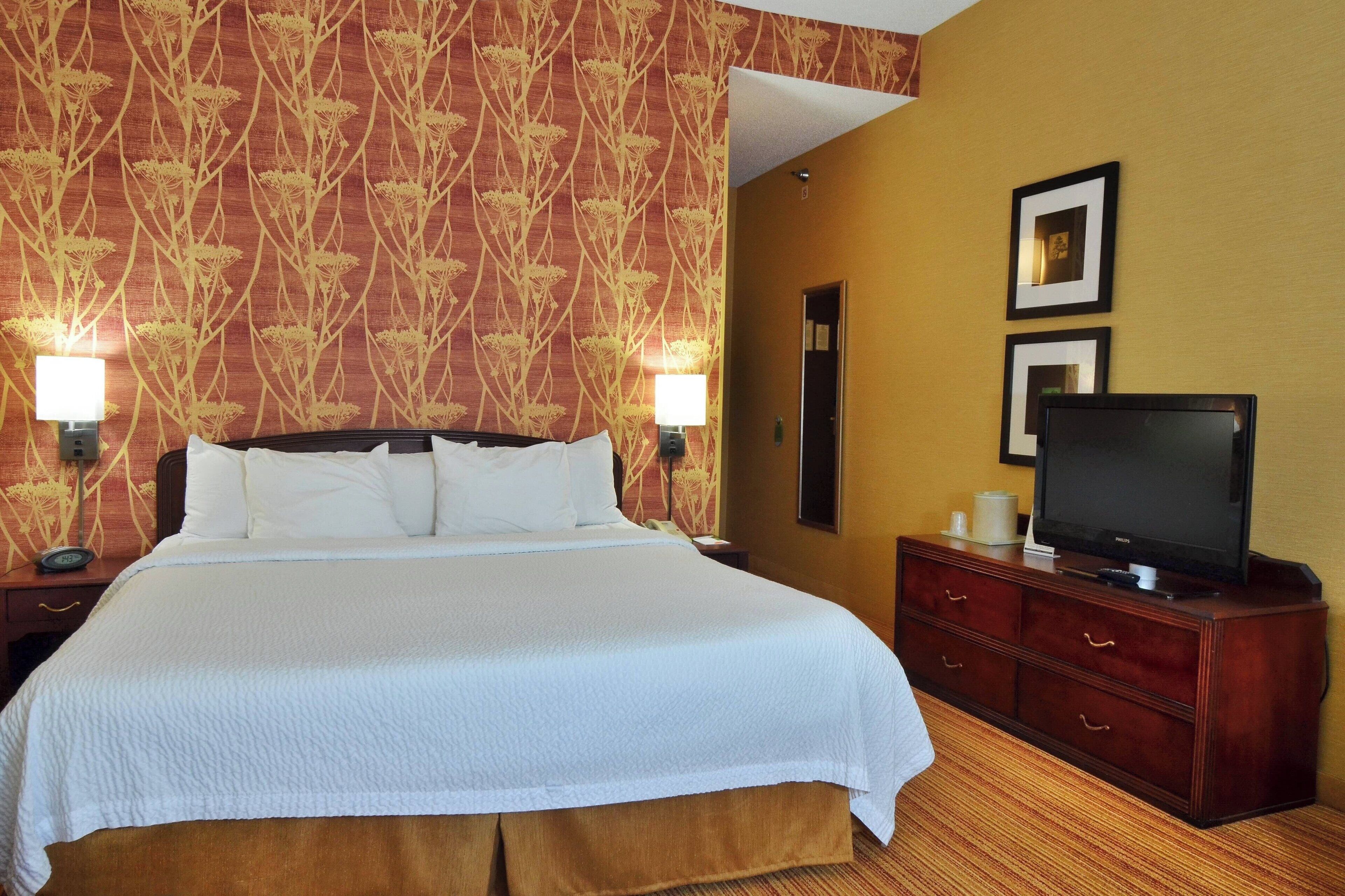 A hotel room with a large bed, a television on a wooden dresser, and two wall-mounted lamps.