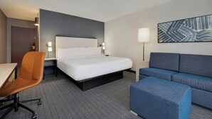 A hotel room with a bed, a desk, a chair, a sofa, and a wall-mounted lamp. - Courtyard by Marriott Indianapolis South (Indianapolis)