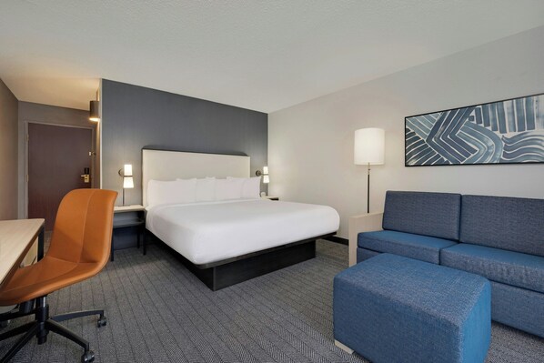 A hotel room with a bed, a desk, a chair, a sofa, and a wall-mounted lamp. - Courtyard by Marriott Indianapolis South (Indianapolis)