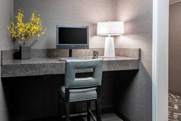 Une chambre d’hôtel moderne avec un bureau en granit foncé, un écran d’ordinateur, un clavier, une chaise, un vase avec des fleurs jaunes et une lampe blanche.