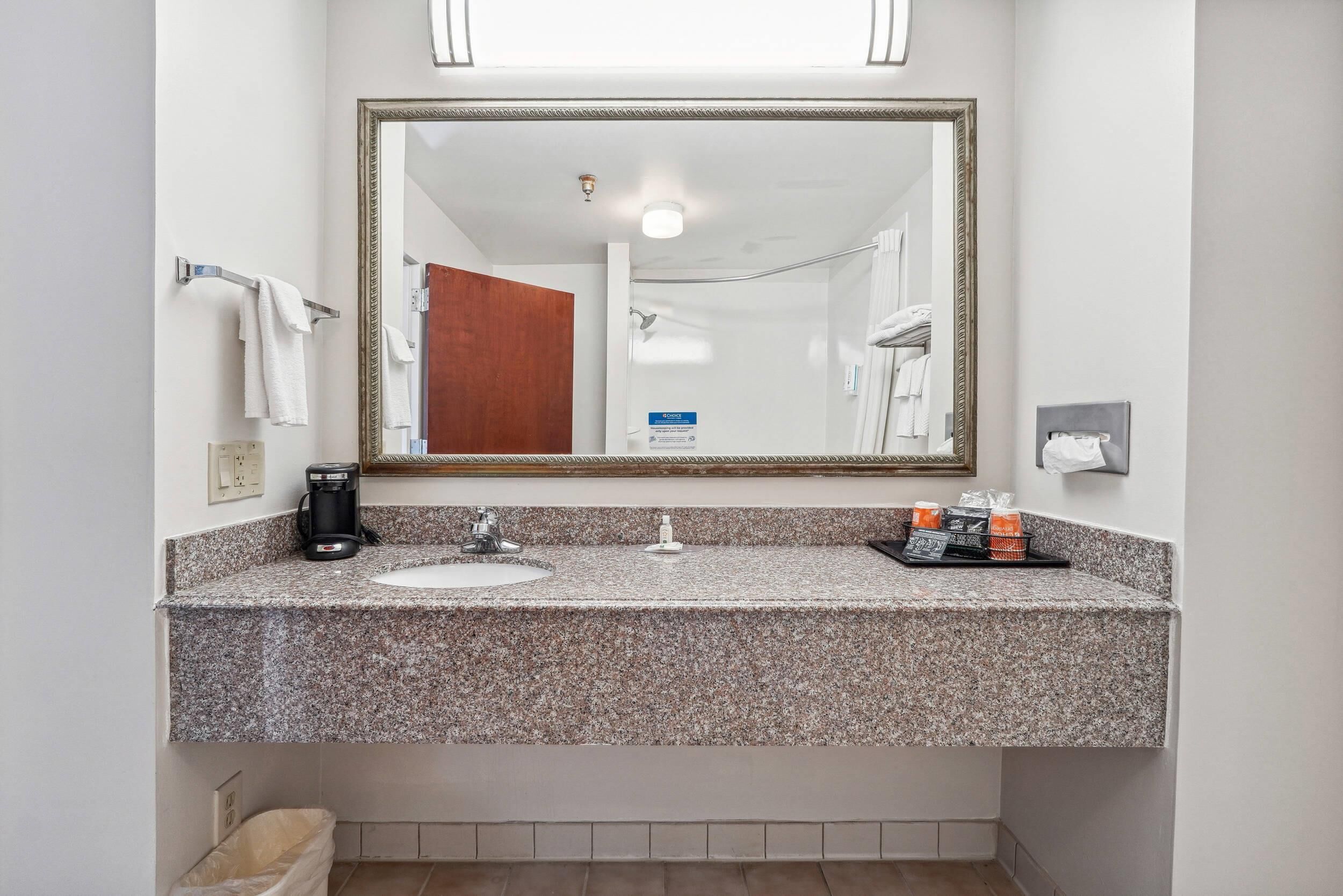 A bathroom with a granite countertop, a sink, a mirror, a towel rack, a coffee maker, and toiletries.