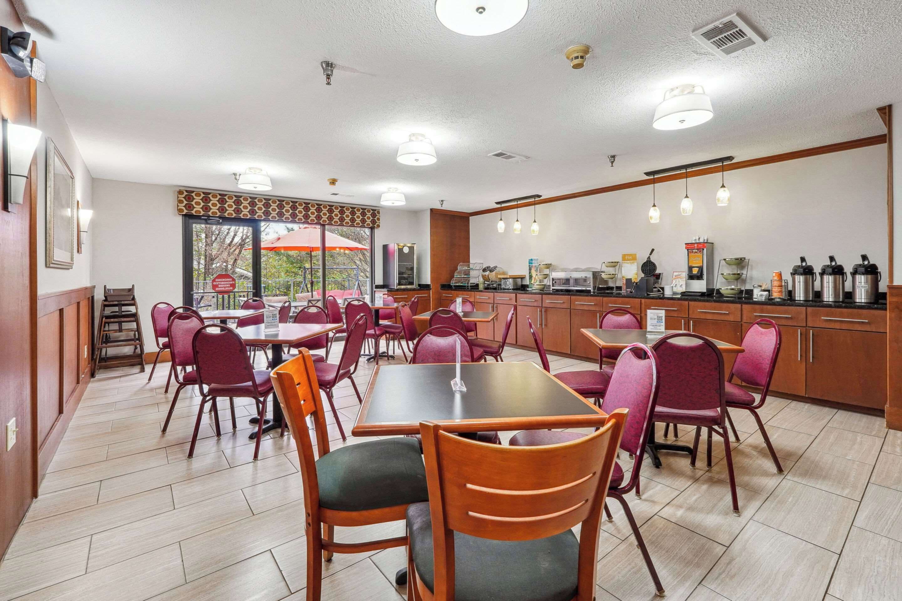 Comedor de hotel con varias mesas y sillas, zona de cocina con máquinas de café y vistas al exterior a través de amplias ventanas.