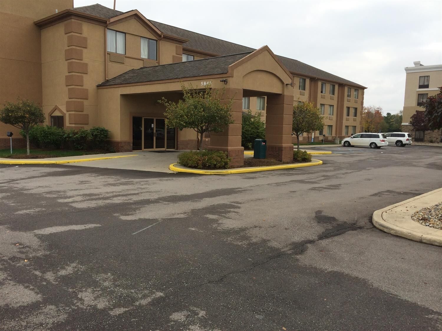 A hotel with a beige exterior, a covered entrance, and a parking lot with two white vehicles.