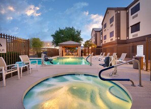 Zona de piscina con sillas de descanso y un jacuzzi, rodeada por una valla y un edificio con varias ventanas.