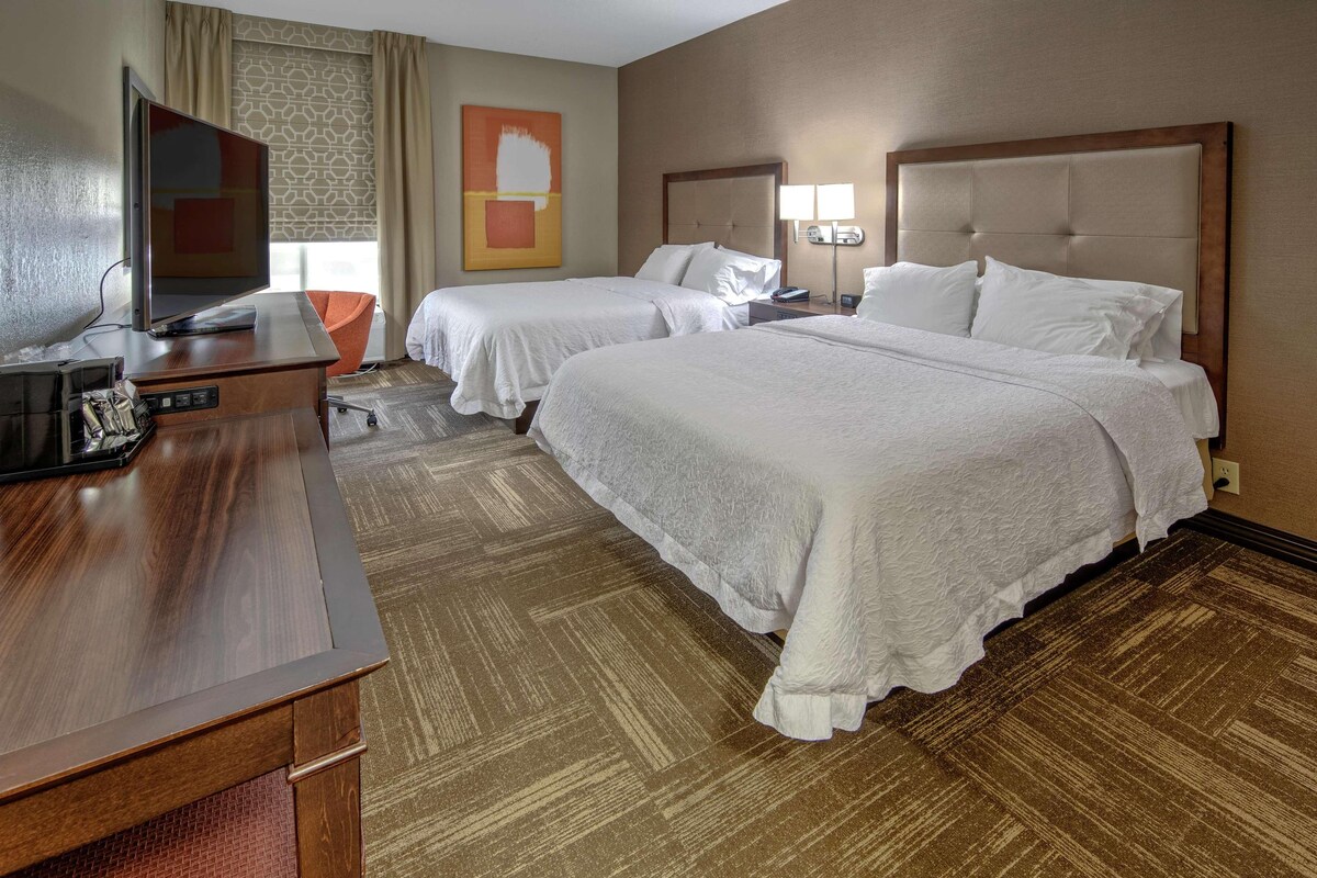 Habitación de hotel con dos camas, un escritorio con televisión de pantalla plana y un ventanal grande con cortinas.