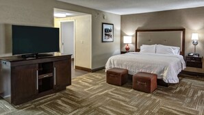 A hotel room with a large bed, a flat-screen TV, a desk with a computer, and a small table with a lamp.