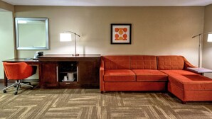 A hotel room with a wooden desk, a red sofa, a chair, a large mirror, a framed picture, and a floor lamp.