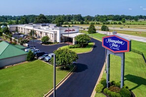A Hampton Inn by Hilton hotel with a parking lot, green lawns, and a clear road in the background.