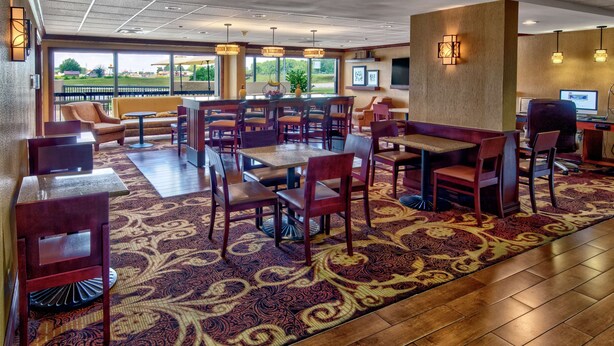 A hotel lobby with a large carpeted area, multiple seating options including tables and chairs, and a view of the outdoors through large windows.
