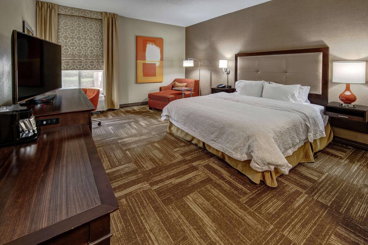A hotel room with a large bed, a desk with a flat-screen TV, a chair, a lamp, and a painting on the wall.