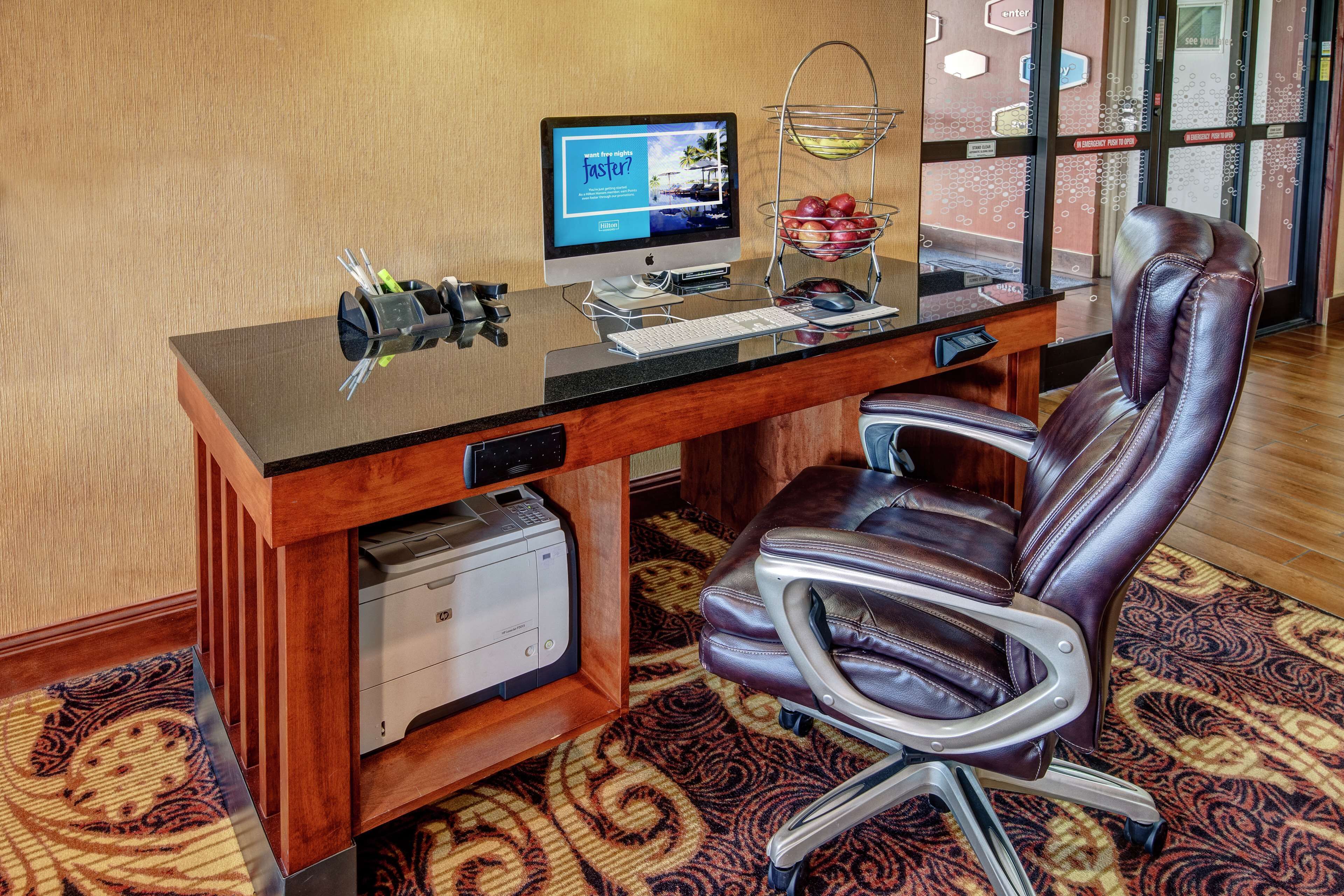 A hotel office with a desktop computer, a printer, a leather chair, and a patterned carpet.