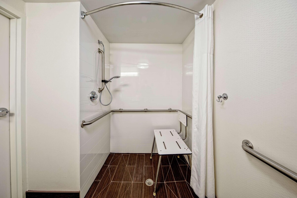 Un baño con paredes blancas, una ducha con barra de apoyo, un pequeño banco blanco y un desagüe en el piso.