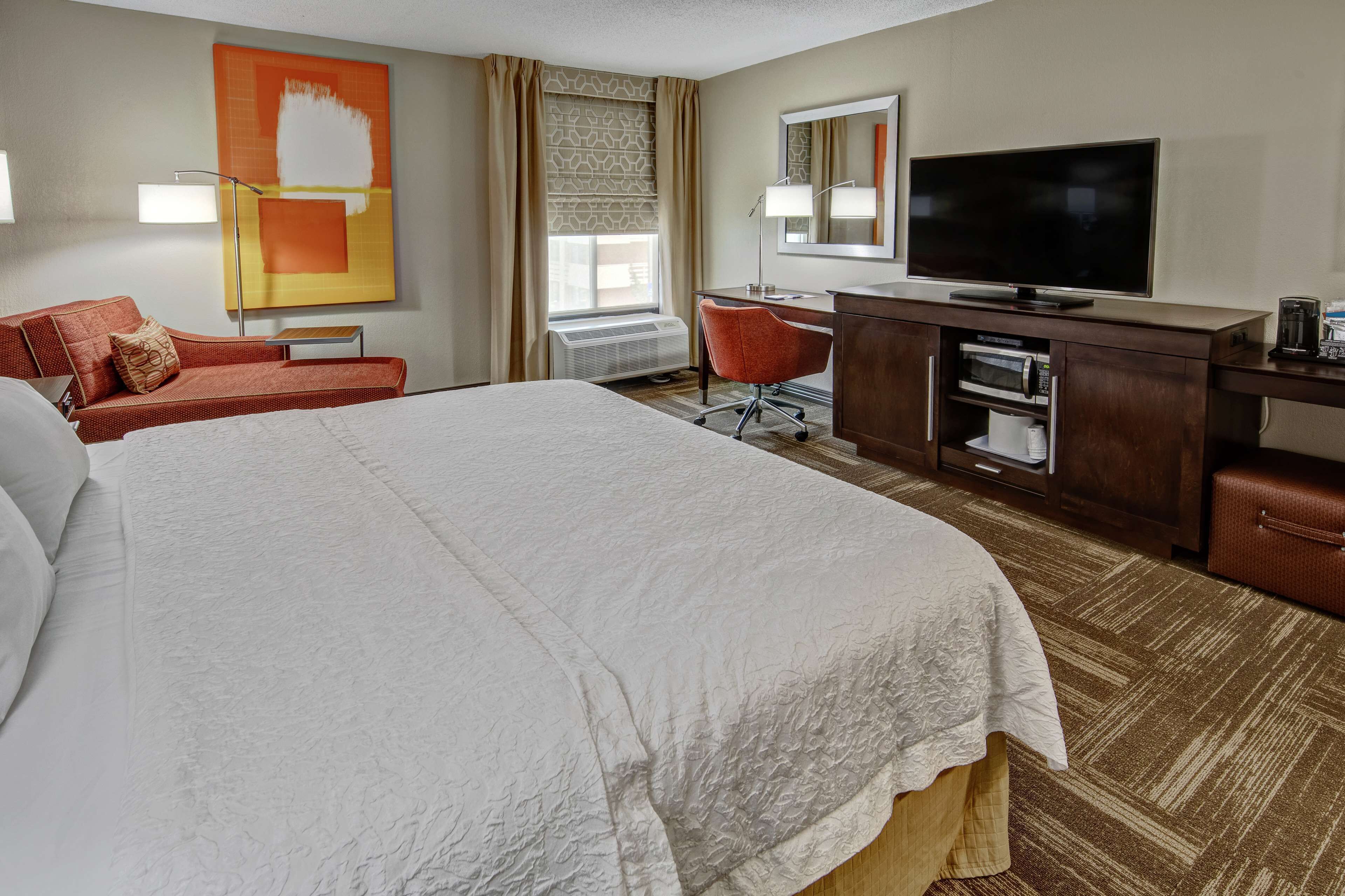 A hotel room with a large bed, a desk with a flat-screen TV, a chair, and a small table.