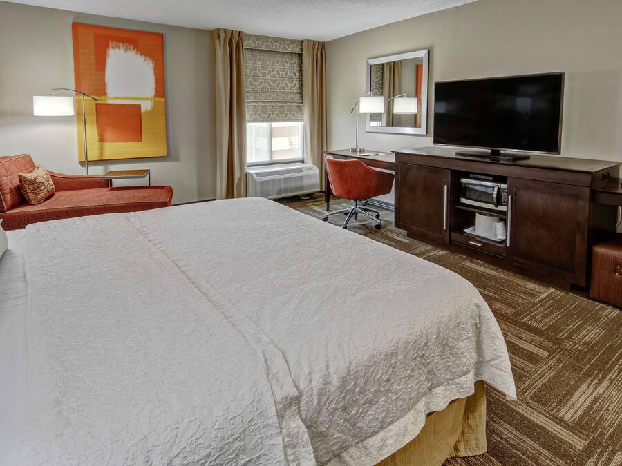 A hotel room with a large bed, a desk with a flat-screen TV, a chair, and a small table.