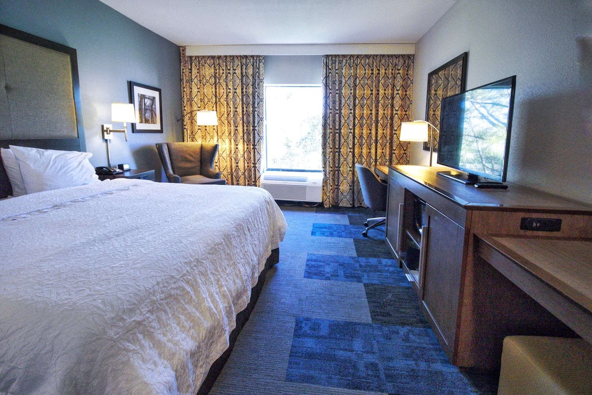 A hotel room with a large bed, a desk with a television, a chair, and a desk lamp.