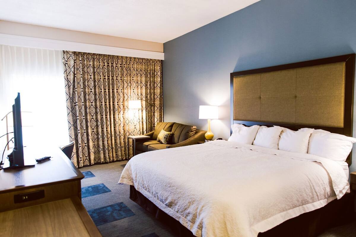 A hotel room with a large bed, a desk with a television, a chair, and a window with curtains.