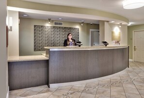 A reception desk with a modern design, a woman standing behind it, and a signboard with hotel shuttle schedule information. - Candlewood Suites Market Center by IHG (Dallas)