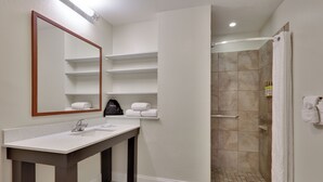 A bathroom with a double sink, a mirror, shelves with towels, and a shower area. - Candlewood Suites Market Center by IHG (Dallas)