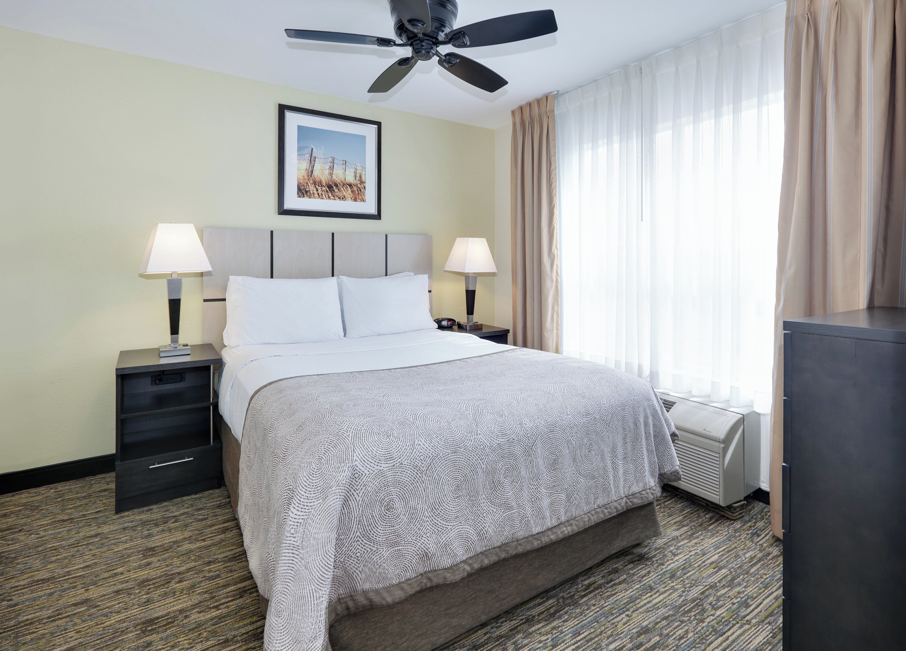 A hotel room with a bed, two bedside tables, a ceiling fan, a framed picture on the wall, and a window with curtains.
