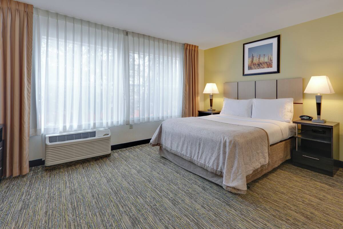 A hotel room with a bed, two bedside tables with lamps, a window with curtains, a radiator, and a picture on the wall.