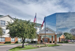 A hotel entrance with a flagpole, a modern glass building, and a tree in the foreground. - Candlewood Suites Market Center by IHG (Dallas)