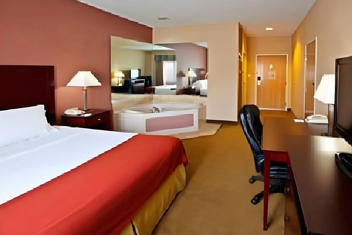 A hotel room with a bed, desk, chair, and a bathroom with a bathtub and mirror.