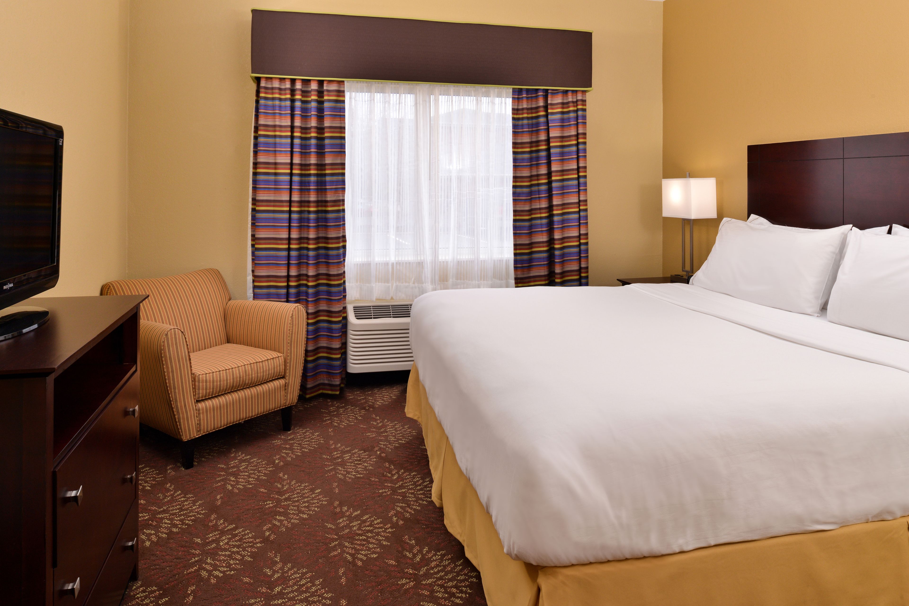 A hotel room with a large bed, a television, a chair, a desk, and a window with curtains.
