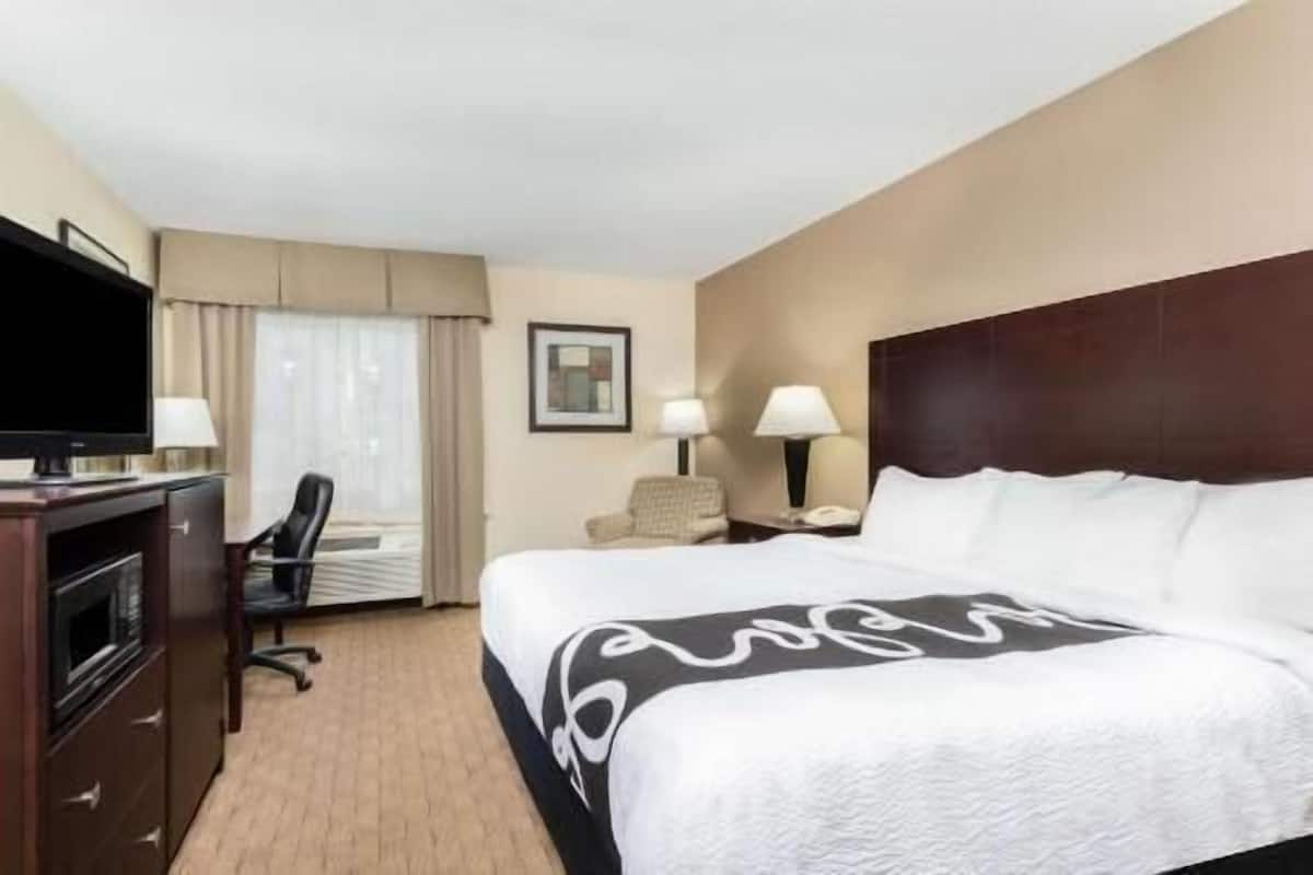 Une chambre d’hôtel avec un lit, un bureau avec une télévision, une chaise, une fenêtre avec des rideaux et un tableau encadré au mur.