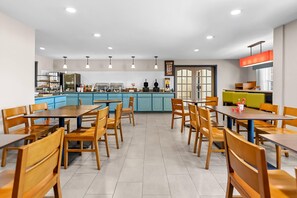 A spacious dining area with wooden tables and chairs, a kitchen in the background, and pendant lighting.