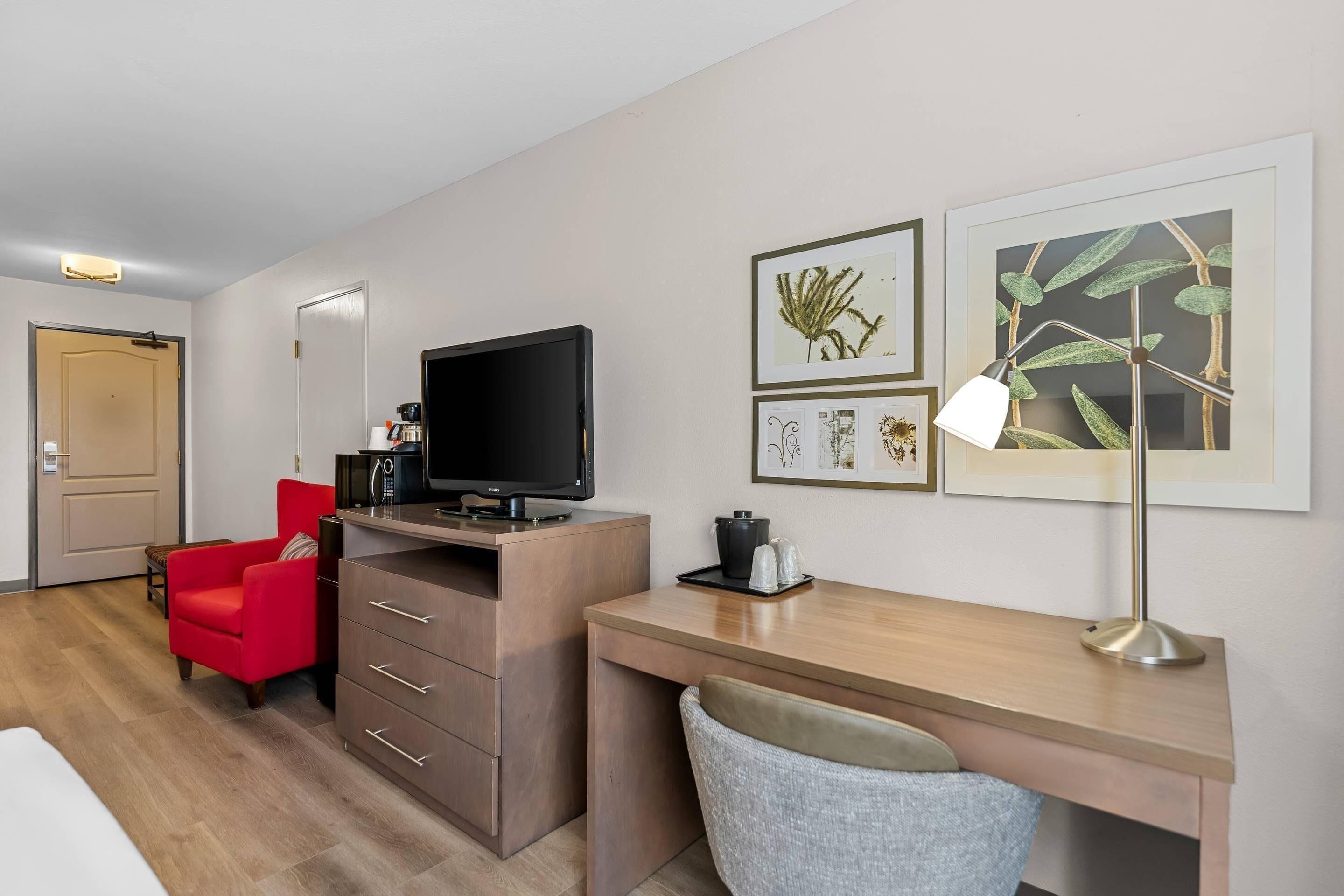A hotel room with a wooden desk, a red chair, a television, and artwork on the wall.