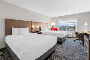 Quarto de hotel com duas camas, uma escrivaninha, uma cadeira, um abajur e uma janela grande.
