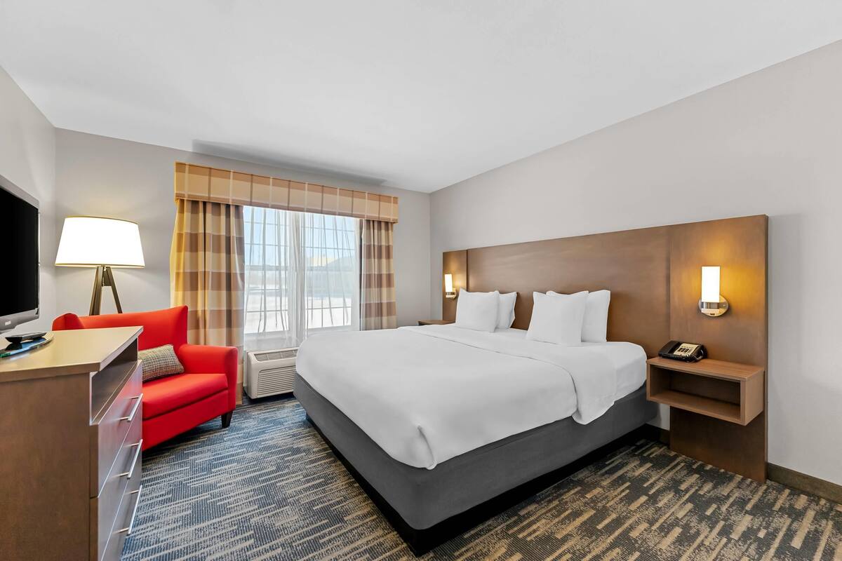 A hotel room with a bed, a desk, a red chair, a TV, a lamp, and a window with curtains.