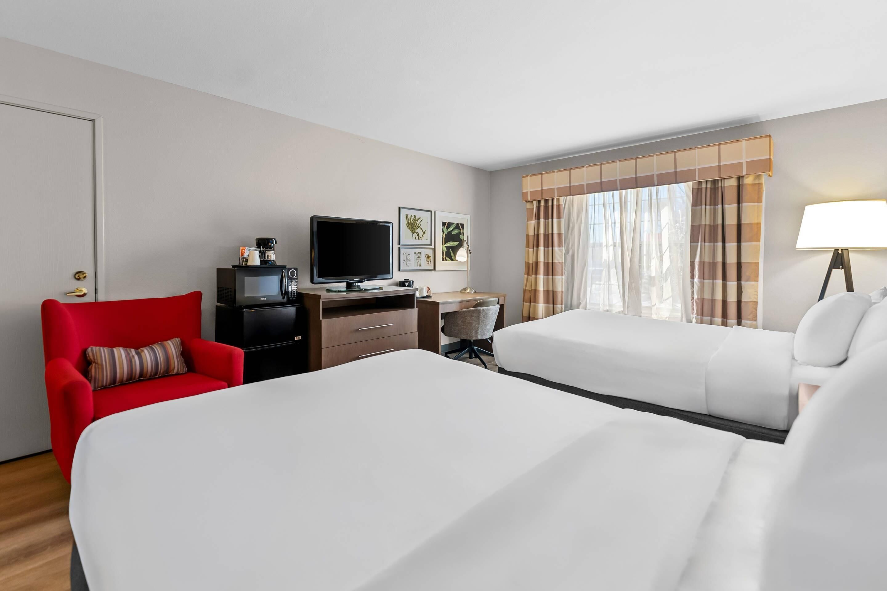 Quarto de hotel com duas camas, uma mesa de trabalho com computador, televisão e uma poltrona vermelha.