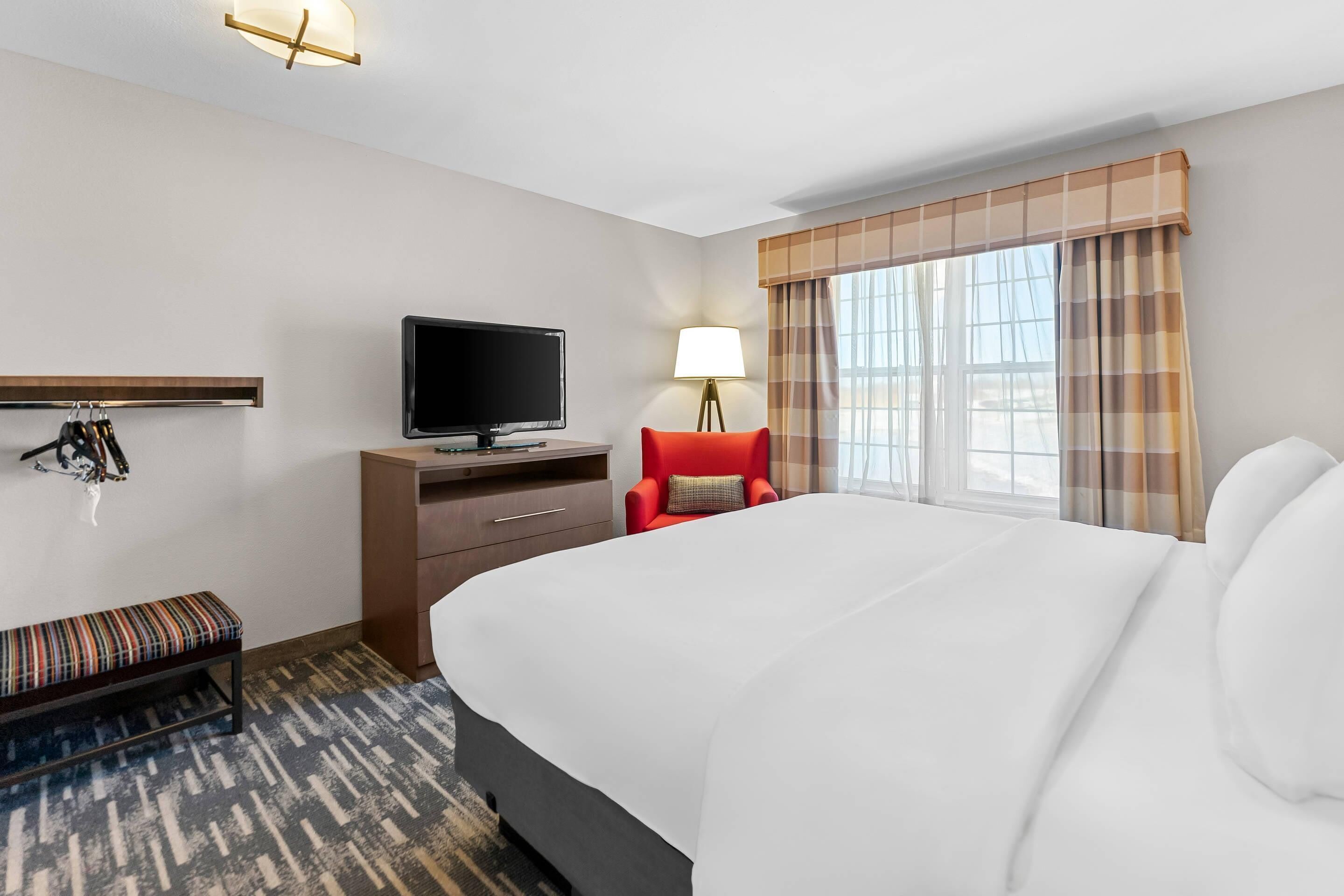 A hotel room with a large bed, a television, a desk with a lamp, a bench, and a window with curtains.