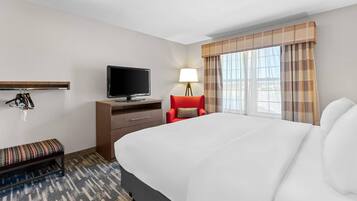 A hotel room with a large bed, a television, a desk with a lamp, a bench, and a window with curtains.
