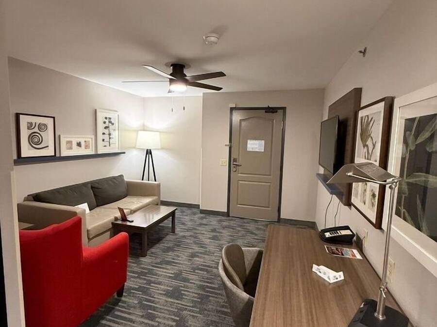 A hotel room with a sofa, a desk, a chair, a lamp, a ceiling fan, and framed pictures on the wall.