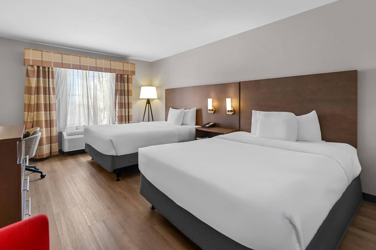 A hotel room with two beds, a desk, a chair, a lamp, and a window with curtains.
