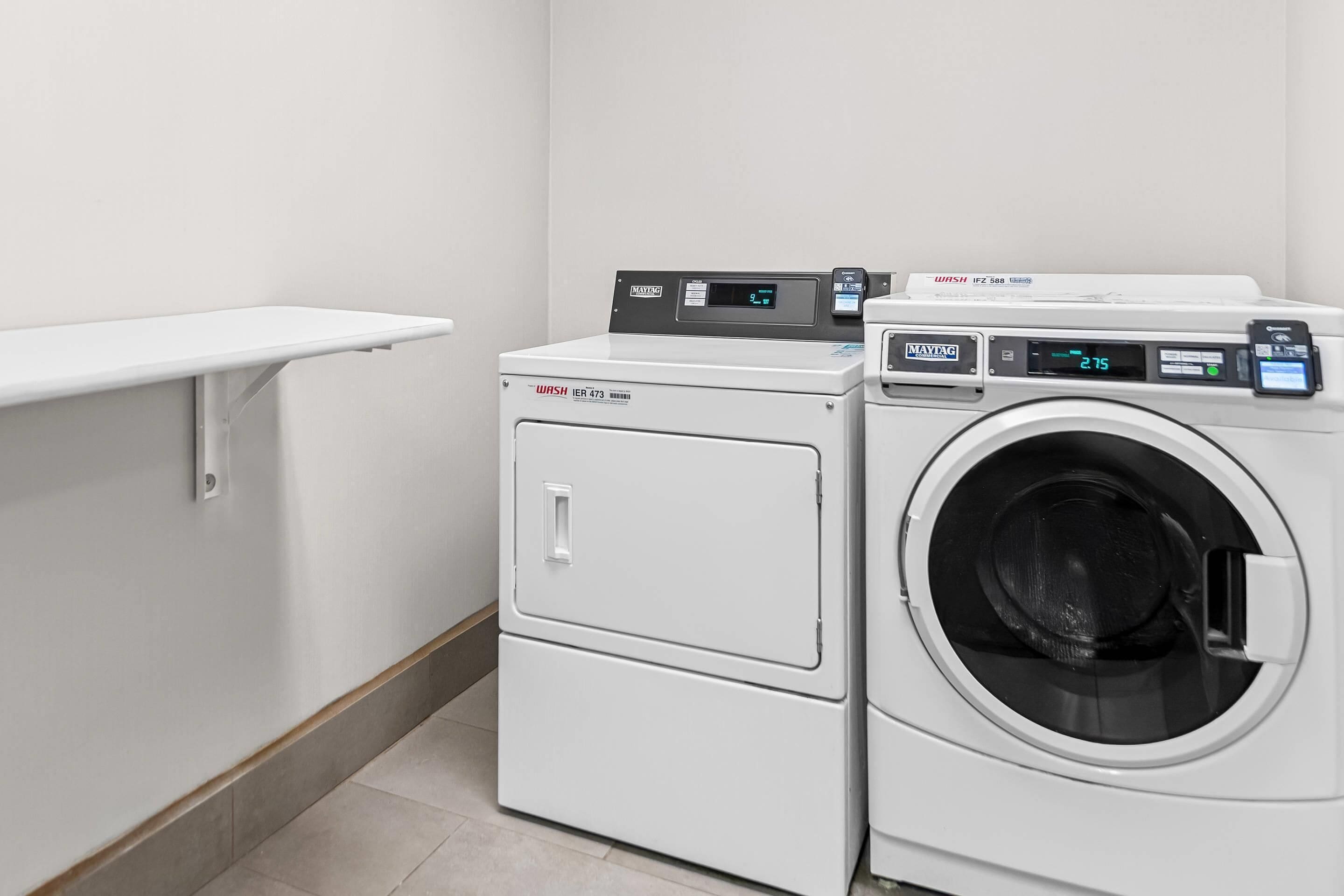 A laundry area with a front-loading washer and dryer, a shelf above the dryer, and a tiled floor.