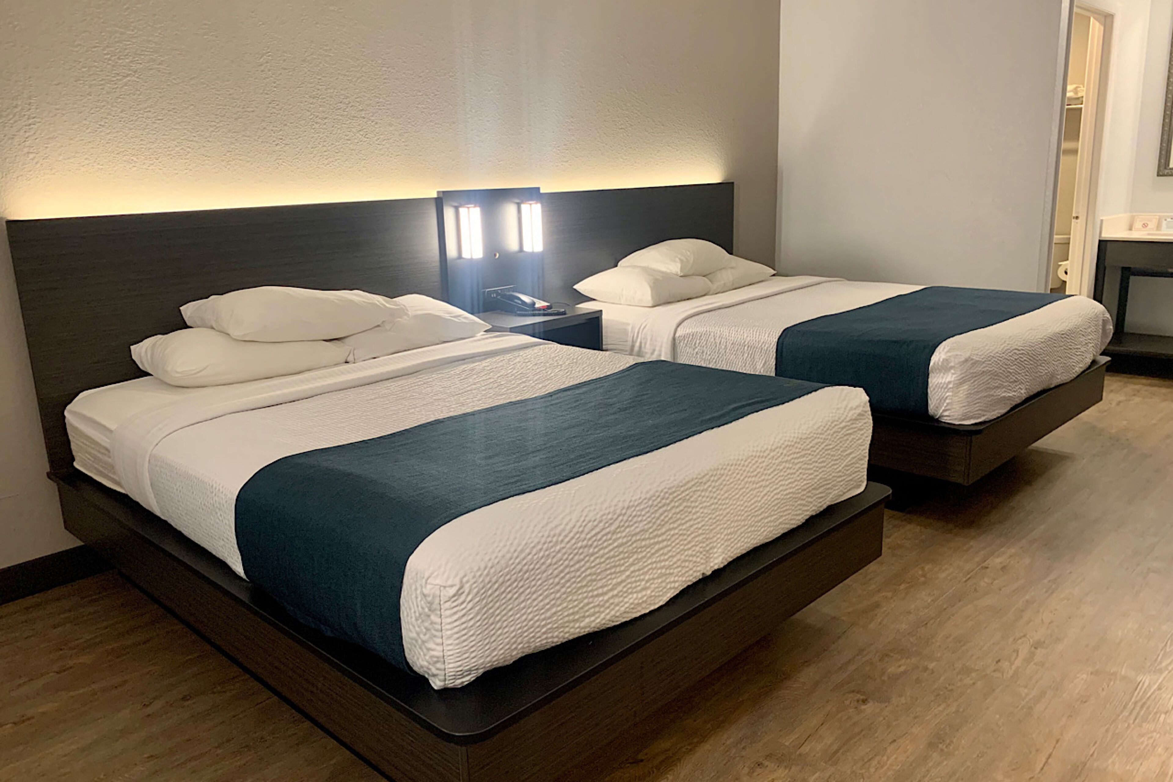 Habitación de hotel con dos camas, cada una con ropa de cama blanca y cabeceros oscuros. Entre las camas, una mesita de noche con lámpara.
