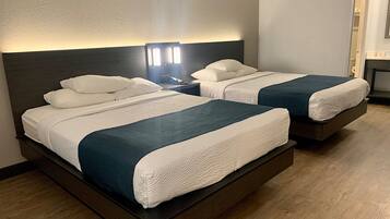 Habitación de hotel con dos camas, cada una con ropa de cama blanca y cabeceros oscuros. Entre las camas, una mesita de noche con lámpara.