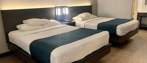 Habitación de hotel con dos camas, cada una con ropa de cama blanca y cabeceros oscuros. Entre las camas, una mesita de noche con lámpara.